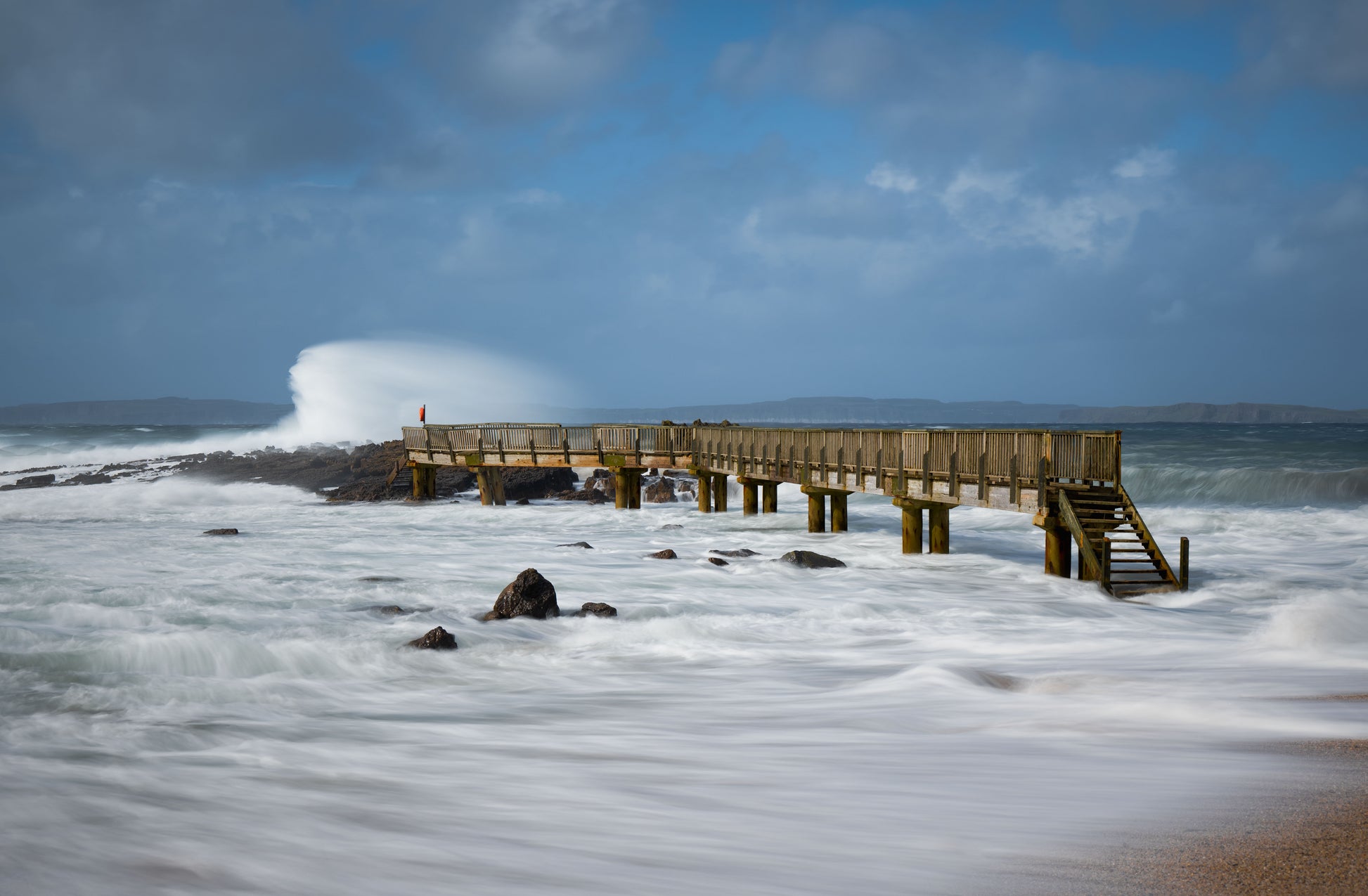 Wooden pier on a rocky beach with waves crashing against it under a blue sky.