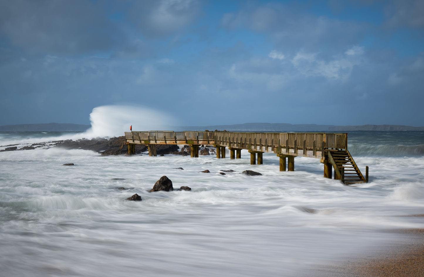 Wooden pier on a rocky beach with waves crashing against it under a blue sky.